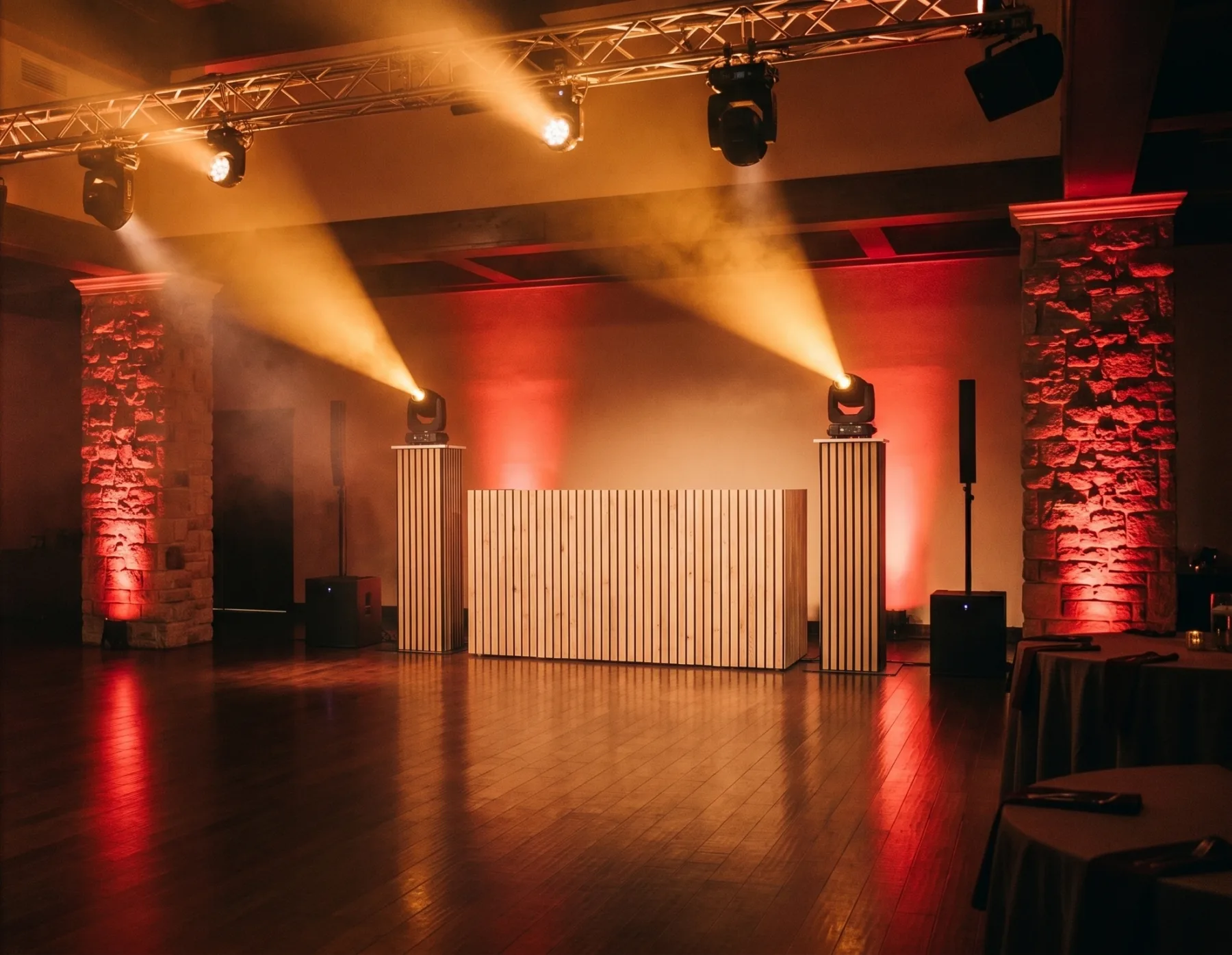 Sala montada antes de una fiesta: cabina de madera, dos torres con cabezas móviles y haces ámbar sobre la pista vacía