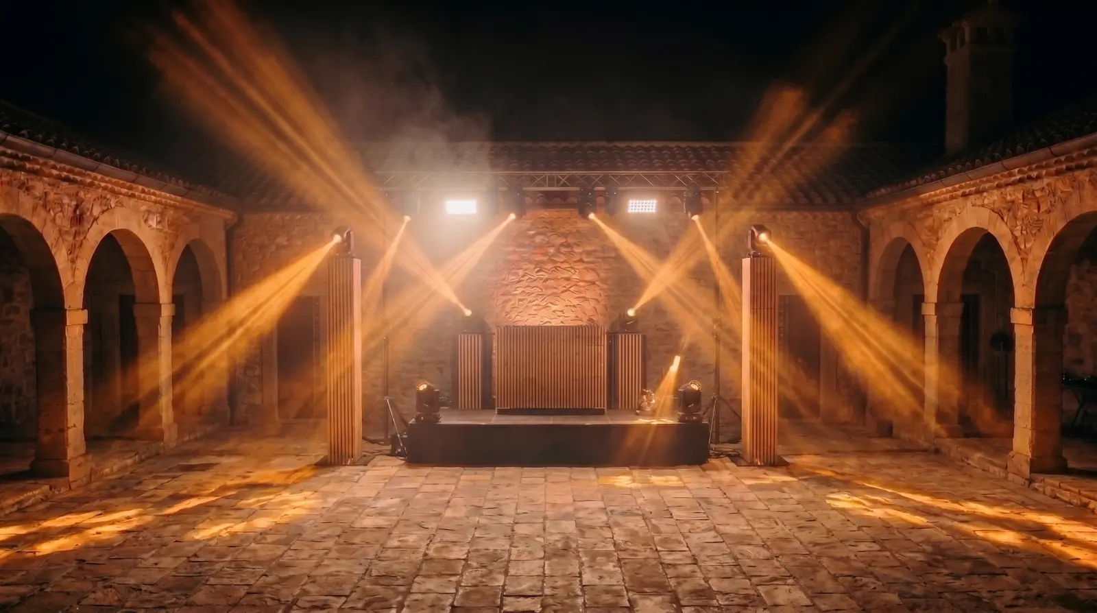 Patio de noche con dos columnas de madera iluminadas a los lados de la cabina y haces ámbar cruzando la pista vacía